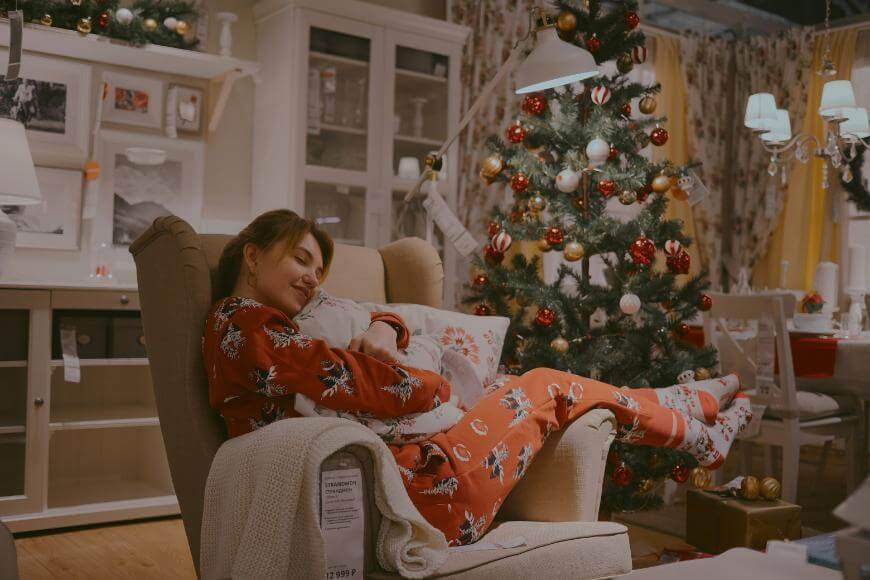 A woman in festive pajamas relaxes in an armchair, hugging a pillow in a cozy, decorated room with a Christmas tree and holiday lights.