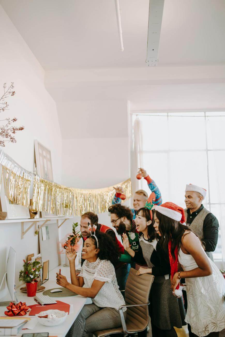 A group of cheerful coworkers wearing festive holiday hats and sweaters gather excitedly around a desk, smiling and celebrating together in a brightly lit office decorated with gold garland.
