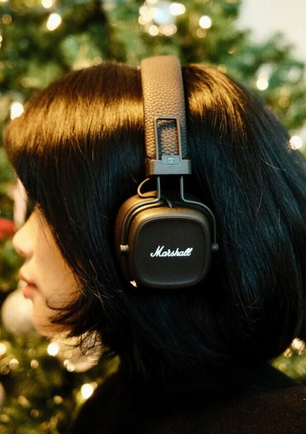 A person with short black hair is wearing black Marshall headphones. There is a decorated, green Christmas tree with lights and ornaments blurred in the background.