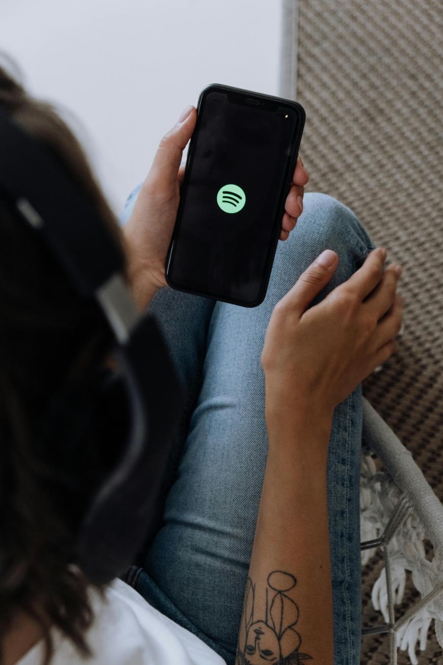 A person wearing headphones holds a smartphone displaying the Spotify logo on the screen. The person is sitting with legs crossed and has a tattoo on their forearm.
