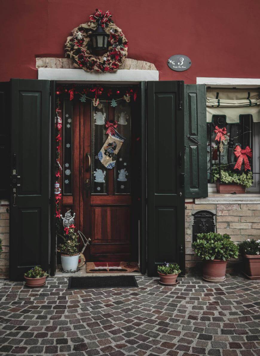 A festive doorway with dark green shutters, a red wall, and a wooden door decorated with a wreath, garland, and Christmas ornaments. Potted plants and a window with red bows add to the holiday decor.