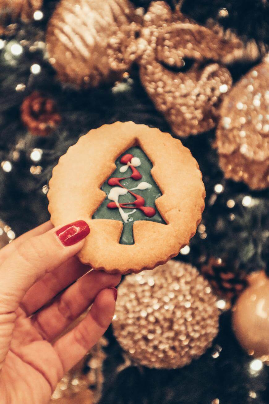 A hand with red nail polish holds a round cookie decorated with a Christmas tree in the center, in front of a decorated Christmas tree with gold ornaments and lights in the background.