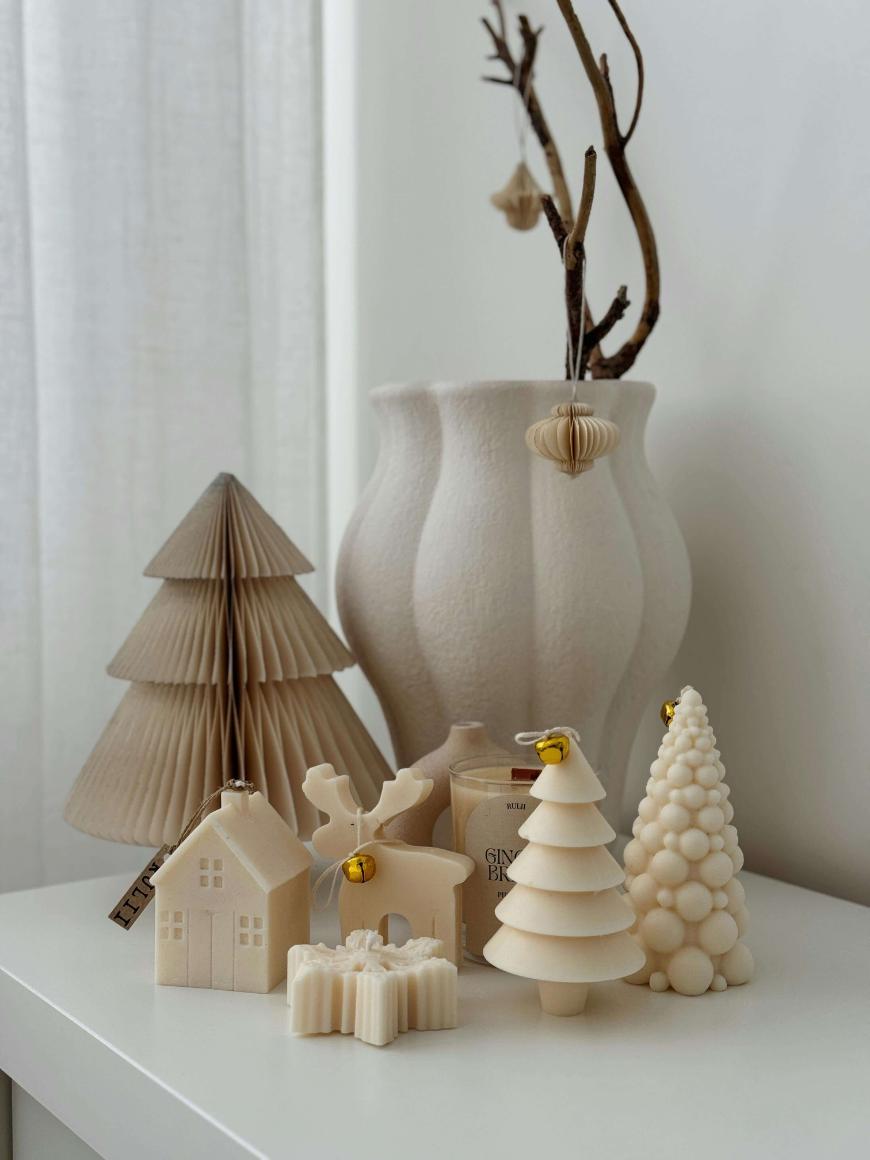 Minimalist holiday decor on a white surface, including cream-colored decorative trees, small house figurines, a gingerbread candle, and a large textured vase with bare branches and paper ornaments.