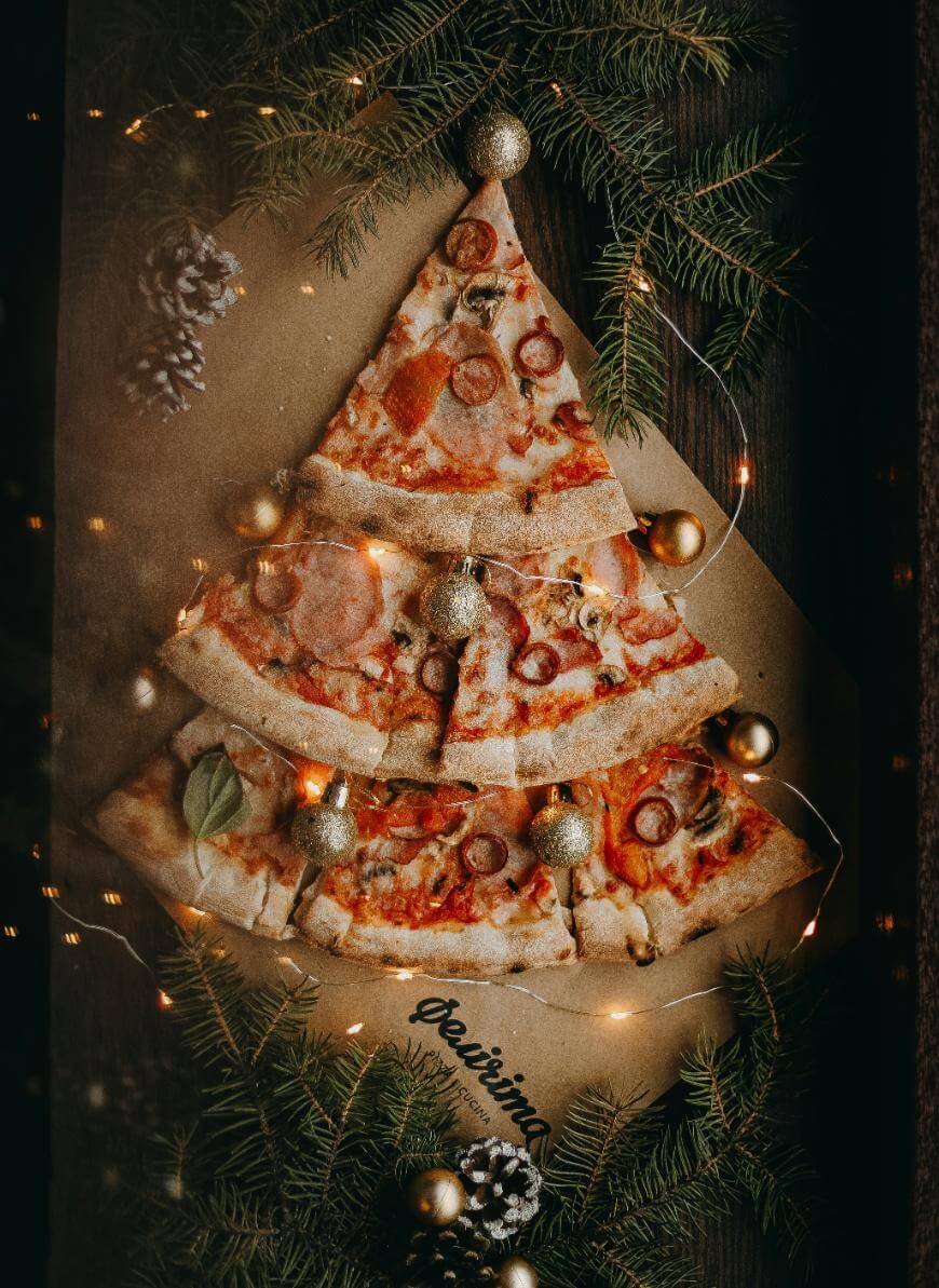 Slices of pizza arranged in the shape of a Christmas tree, decorated with small gold ornaments and surrounded by pine branches, pinecones, and string lights.