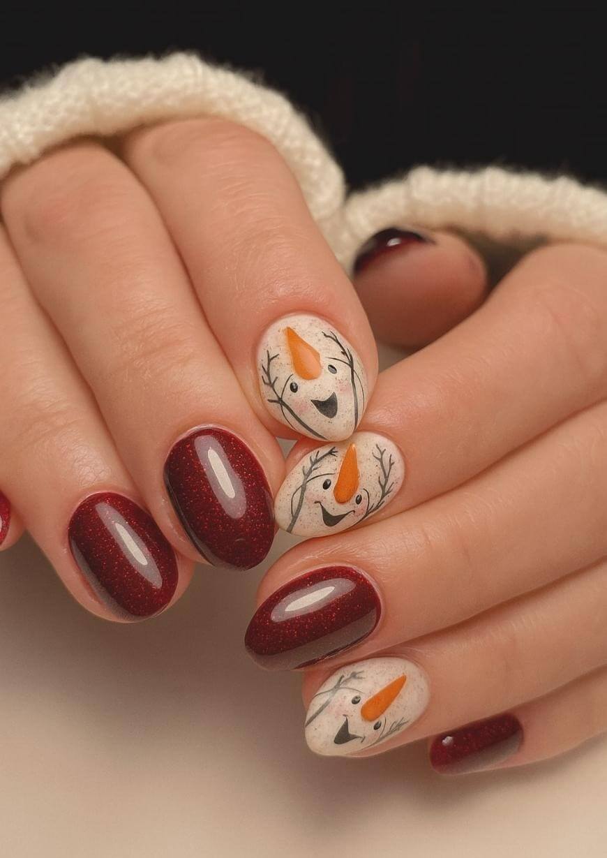 A hand with almond-shaped nails painted dark red, except for two accent nails on each hand. The accent nails feature cute snowman faces with orange carrot noses, black eyes and mouths, and twig-like lines for arms.