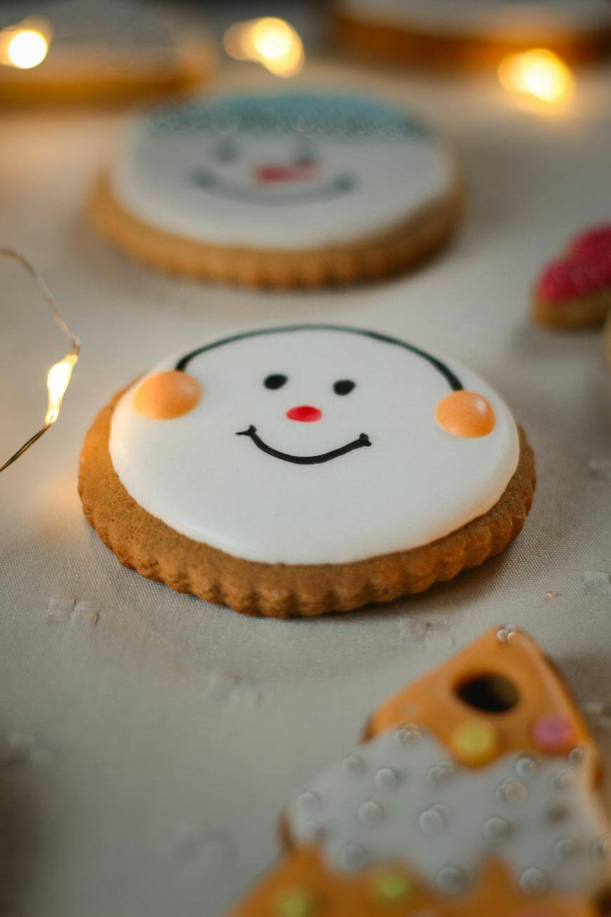 A round cookie decorated with white icing, a smiling face, rosy cheeks, and black earmuffs, surrounded by festive lights and other decorated cookies.