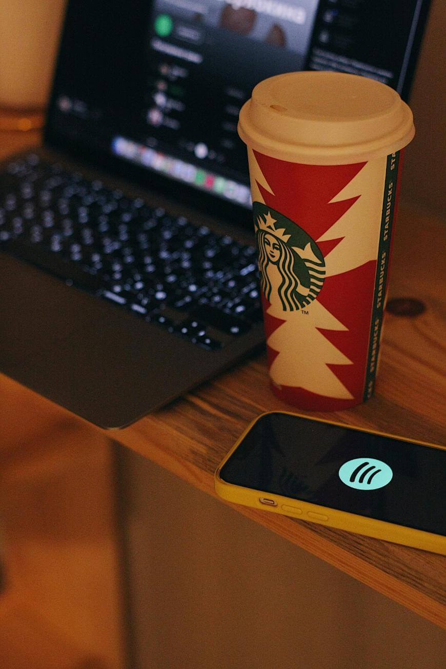 A Starbucks holiday cup sits on a wooden surface next to an open laptop with a blurred screen and a yellow smartphone displaying the Spotify logo.