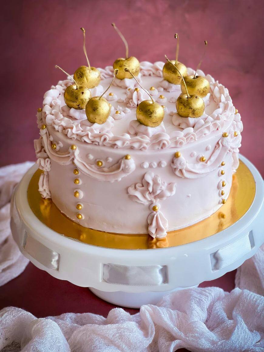 A decorative pink frosted cake topped with gold-painted cherries sits on a white and gold cake stand against a soft pink background. Delicate icing details and gold pearls adorn the sides.