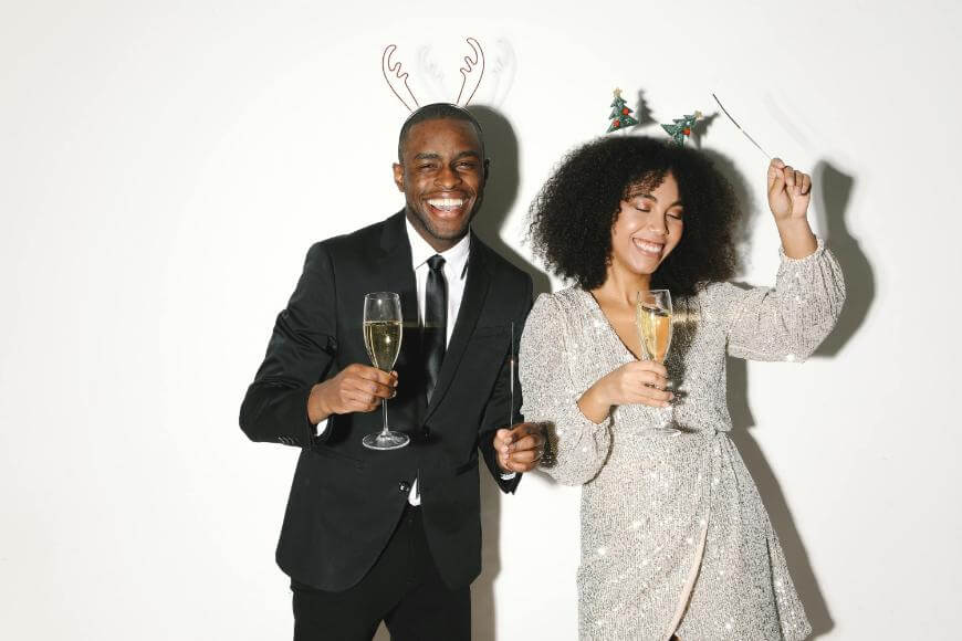 A man in a suit and a woman in a sparkly dress, both wearing festive headbands, smile and hold champagne glasses while dancing at a celebration.