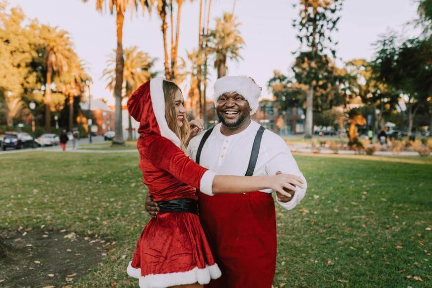 A man in a Santa hat and red suspenders smiles while dancing with a woman dressed in a red Santa dress and hood in a sunny park with palm trees in the background.