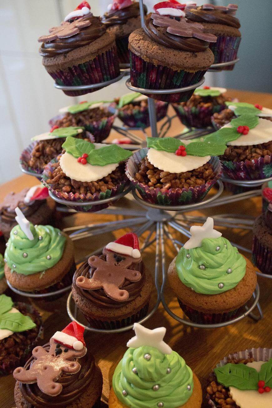 A tiered stand of festive cupcakes decorated with green frosting, fondant holly leaves, gingerbread men, stars, and Santa hats, arranged on a wooden surface.