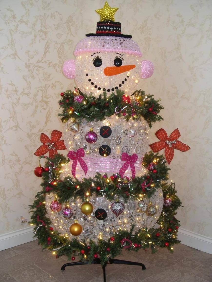 A festive snowman decoration made from clear plastic cups, adorned with ornaments, garlands, and lights, with a pink hat, orange carrot nose, and a star on top, standing against a light wall.