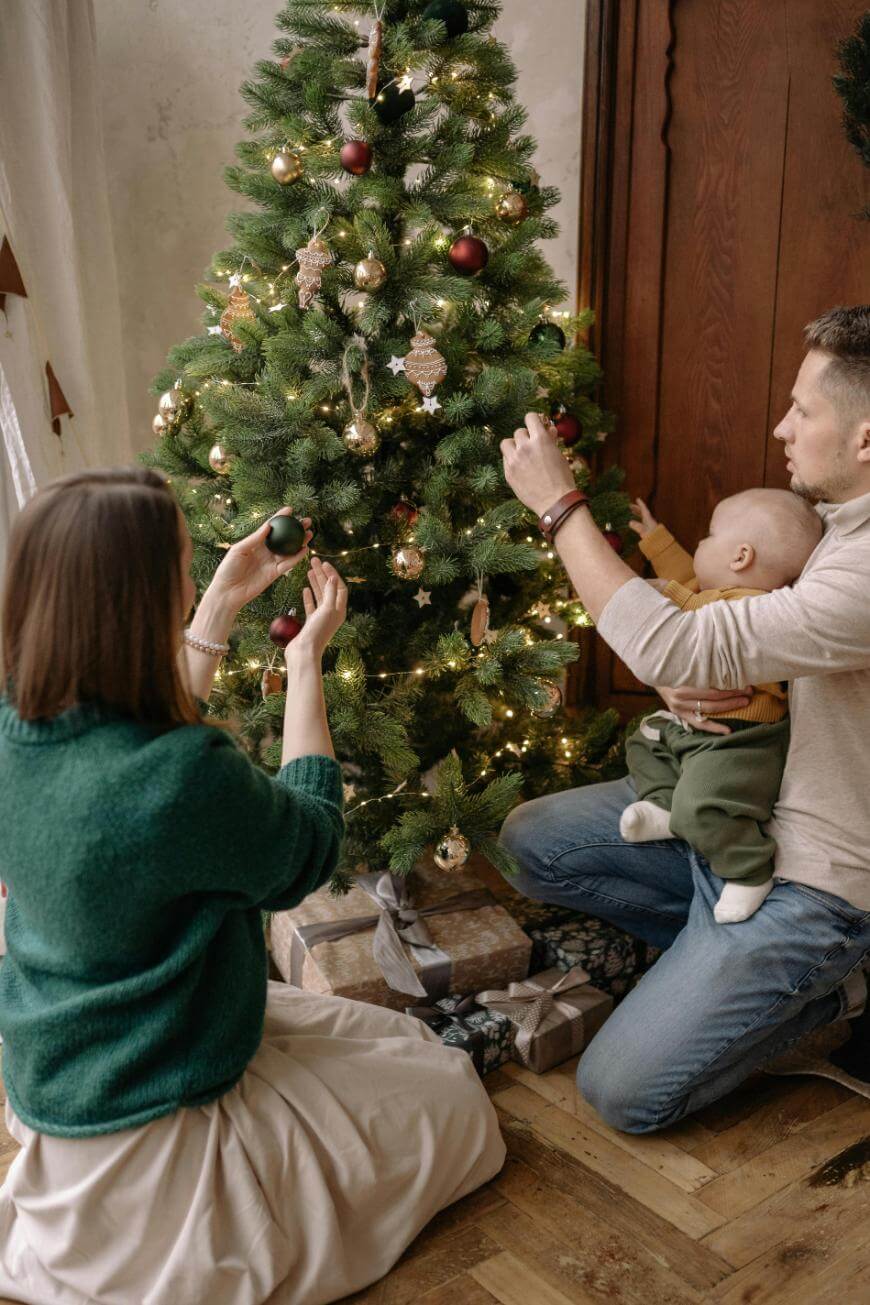 A family decorates a Christmas tree together. An adult holds a baby while placing an ornament, and another adult kneels nearby, hanging a bauble. Wrapped presents sit under the tree.