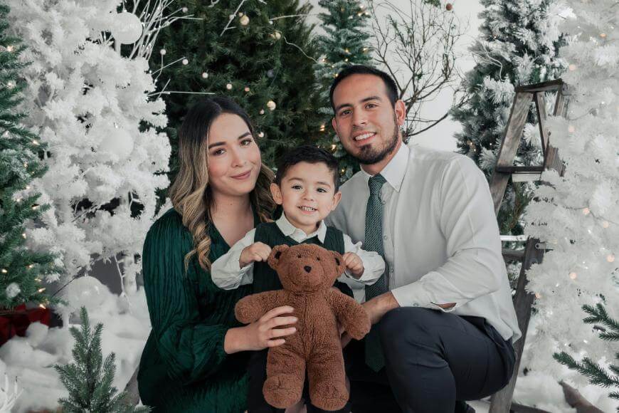 A family of three poses for a holiday photo surrounded by white and green Christmas trees. The parents are kneeling on either side of their young son, who is holding a brown teddy bear and smiling at the camera.