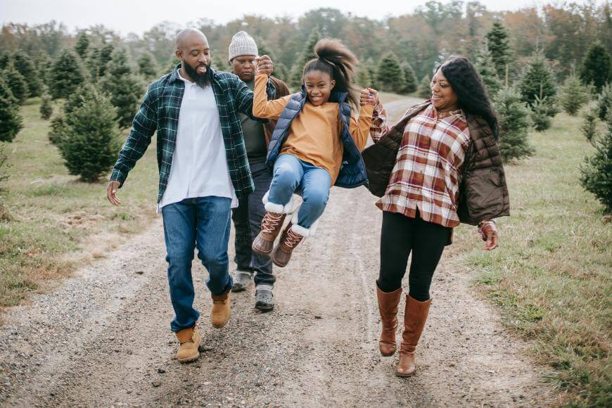 Four people walk along a dirt path in a tree farm. Two adults swing a smiling child between them, while another adult walks behind. They all wear fall clothing and look happy outdoors.