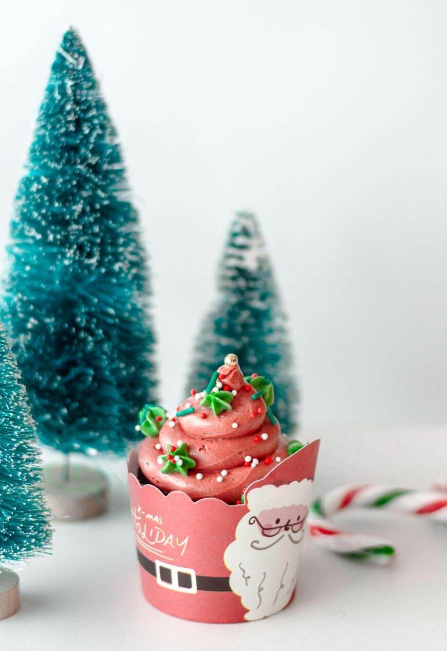 A festive cupcake with pink frosting, green and white star sprinkles, and a paper Santa wrapper sits in front of small blue Christmas trees. A candy cane is in the background on a white surface.