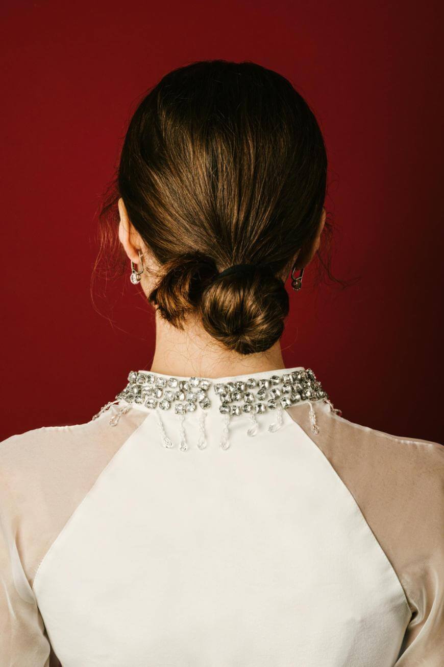 A woman with brown hair styled in a low bun, wearing a white top with a beaded neckline, is shown from behind against a deep red background.