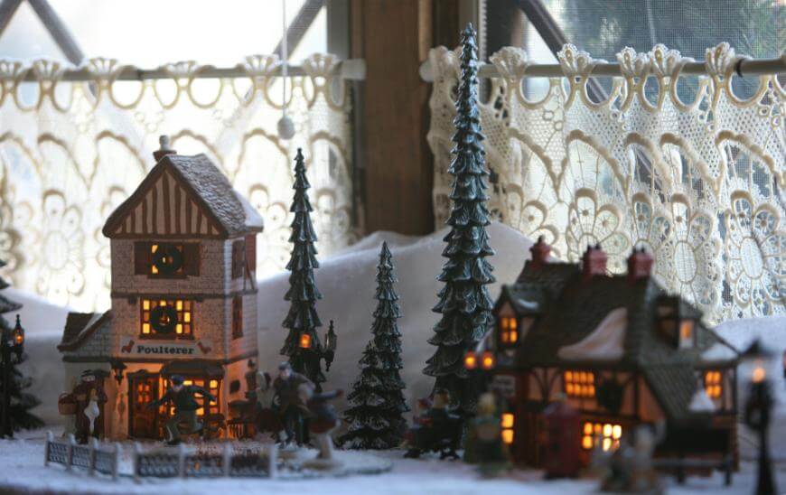 A miniature winter village scene with snow-covered houses, pine trees, and warm yellow lights inside the buildings, set against a window with lace curtains.