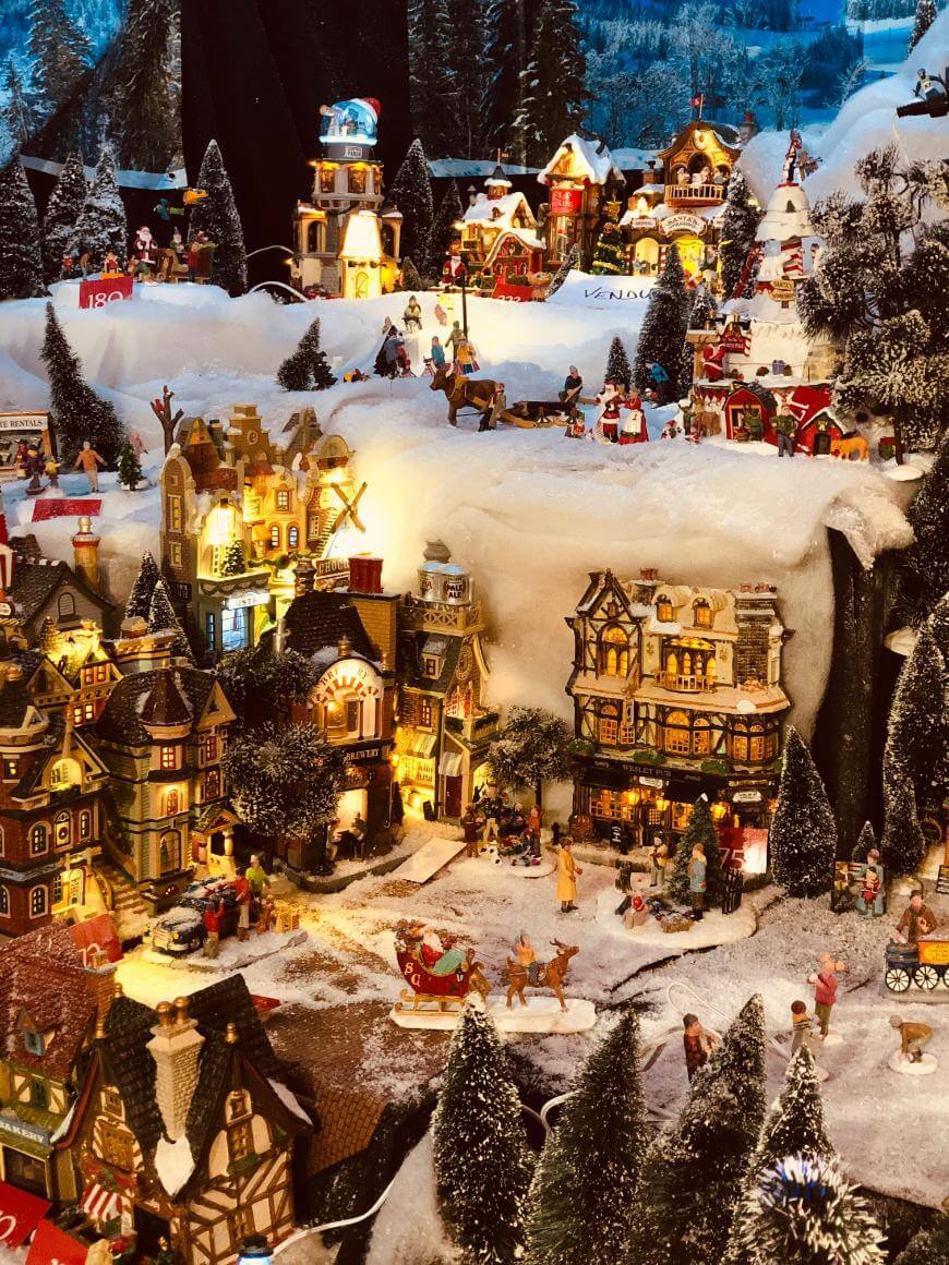 A miniature winter village scene with snow-covered buildings, tiny figurines, pine trees, and festive lights, creating a charming holiday atmosphere. Some villagers walk, skate, and gather in the snowy streets.