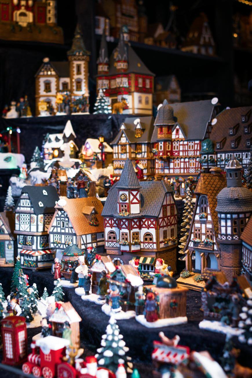 Miniature model village scene featuring detailed, colorful half-timbered houses, tiny trees, festive decorations, and small figurines, creating a lively, winter holiday atmosphere.