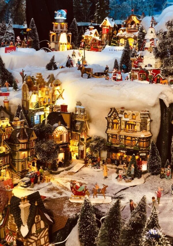 A detailed miniature winter village scene with snow-covered buildings, glowing lights, tiny figurines, trees, and a sleigh, creating a festive holiday atmosphere.