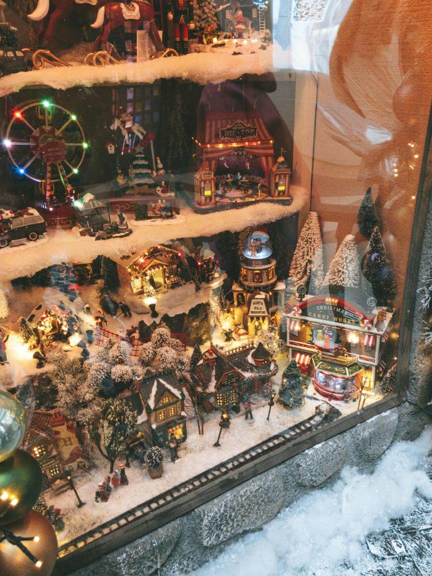 A festive shop window display features a miniature snowy Christmas village with twinkling lights, tiny houses, trees, a Ferris wheel, and various holiday figures, all surrounded by artificial snow.