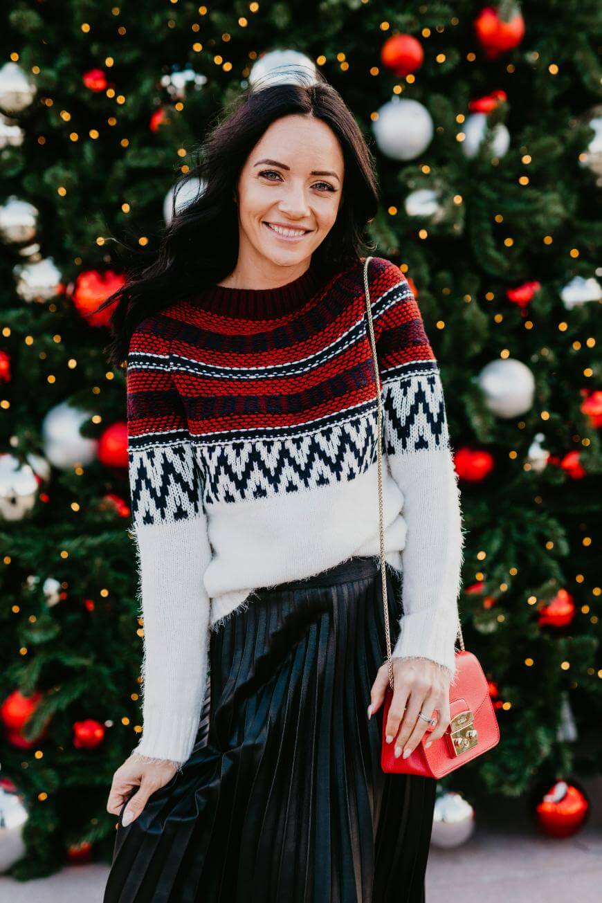 A woman with long dark hair stands in front of a decorated Christmas tree, wearing a festive red, white, and black sweater, a black pleated skirt, and holding a small red purse.