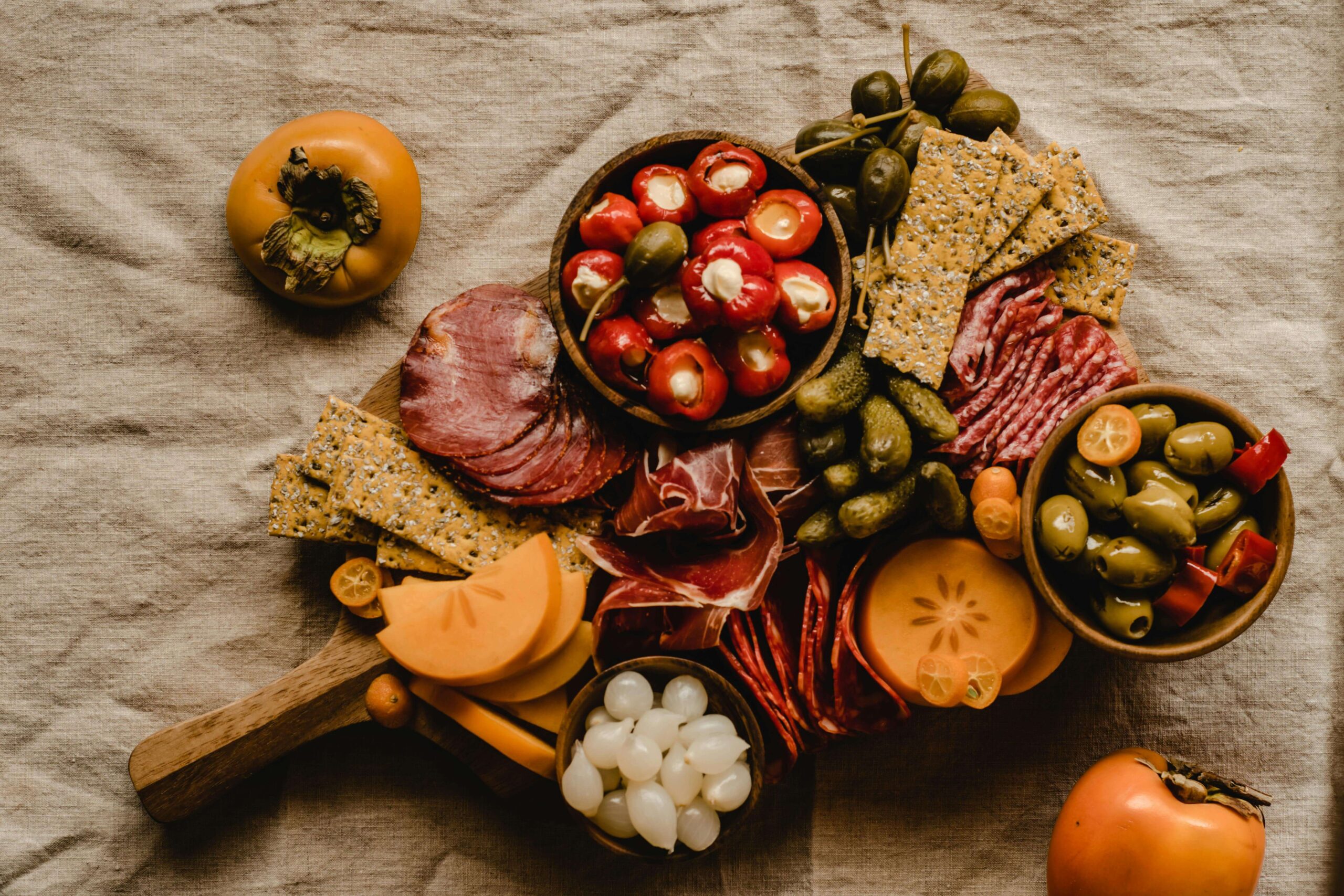 A charcuterie board with crackers, sliced meats, stuffed peppers, olives, pickles, pearl onions, and slices of persimmon, set on a beige cloth with whole persimmons beside the board.