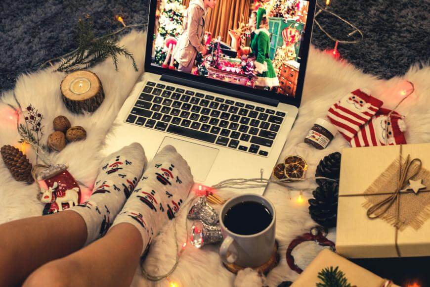 A person wearing festive socks sits on a white rug with a cup of coffee, watching a Christmas movie on a laptop, surrounded by holiday decorations, nuts, wrapped gifts, and string lights.
