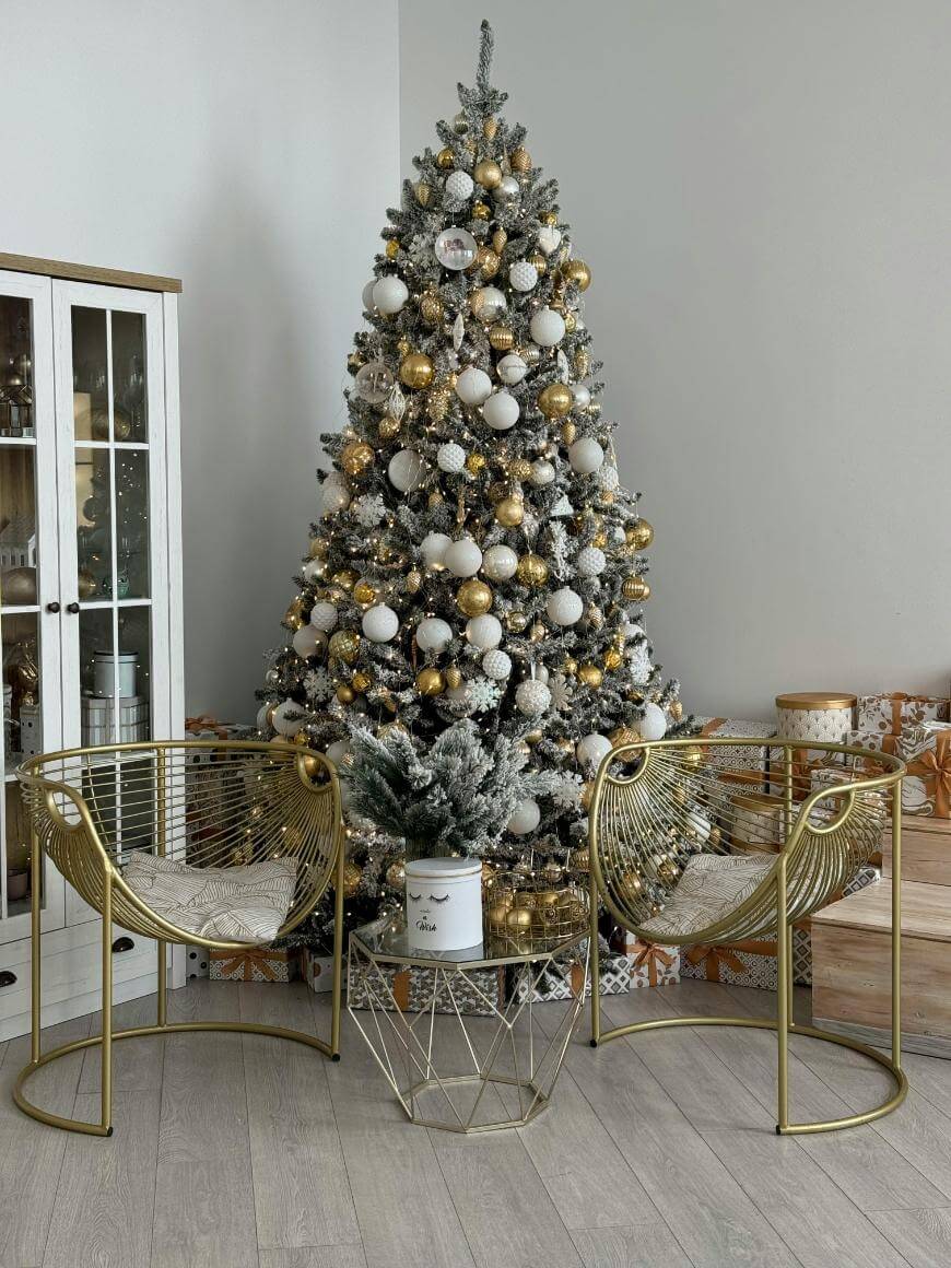 A Christmas tree decorated with gold and white ornaments stands in a corner. Two modern gold chairs and a small table with a candle are in front of it. Presents are stacked on the right, and a cabinet is on the left.