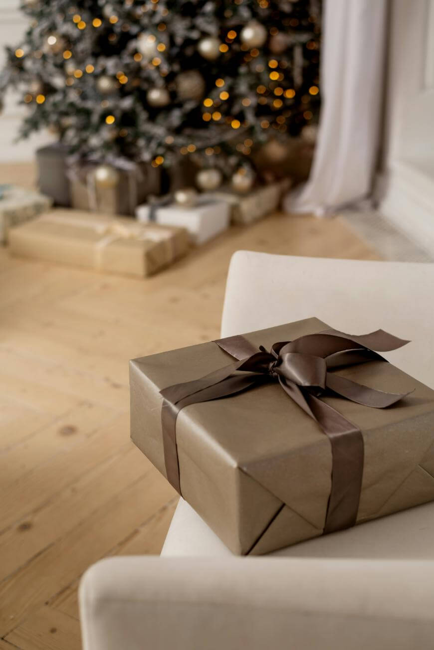 A neatly wrapped gift with brown paper and a dark brown ribbon sits on a white chair, with a decorated Christmas tree and more presents in the background.