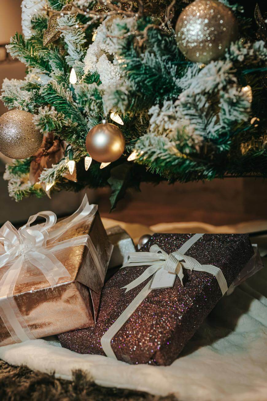 Wrapped gifts with white ribbons are placed under a decorated Christmas tree with golden ornaments, frosted branches, and warm white string lights.
