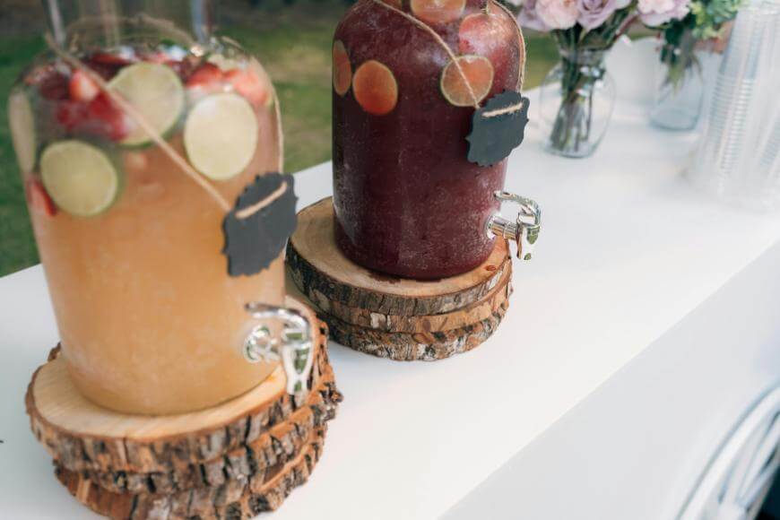 Two large glass drink dispensers sit on rustic wooden slabs. One dispenser holds a yellow beverage with lime and strawberries; the other has a dark red drink with fruit slices. Both have spigots and blank chalkboard labels.