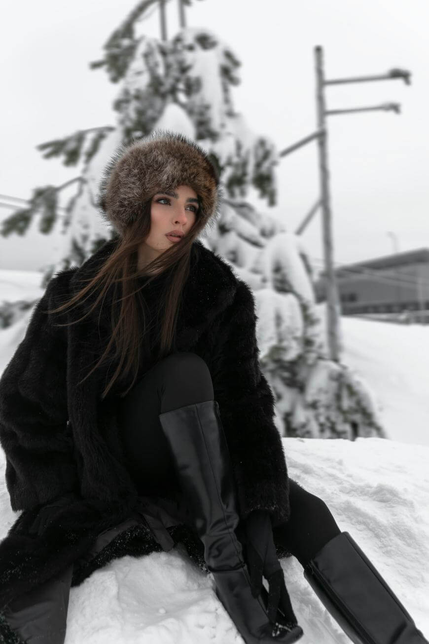 A woman wearing a fur hat, black coat, and tall black boots sits on snow, with snow-covered trees and a building in the background on a cloudy winter day.