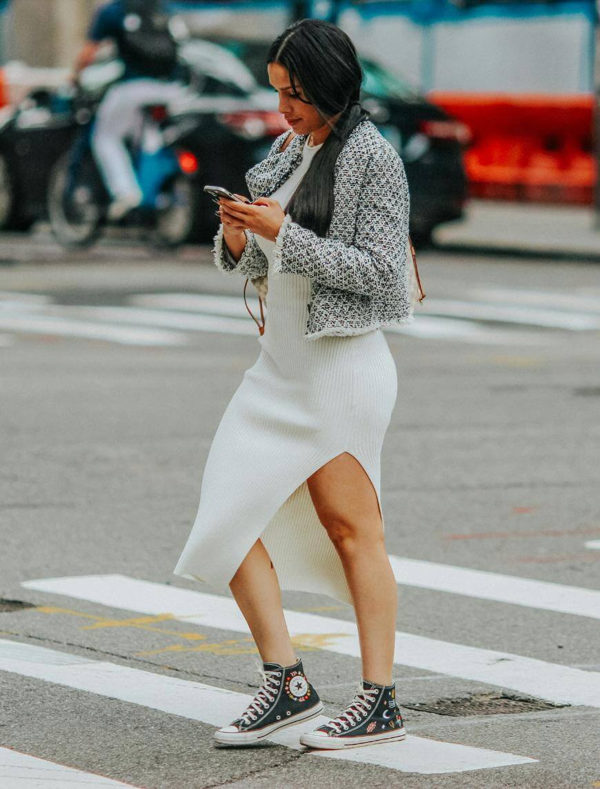 A woman with long dark hair, wearing a white ribbed dress with a side slit, a cropped patterned jacket, and black high-top Converse sneakers, walks across a city street while looking at her phone.