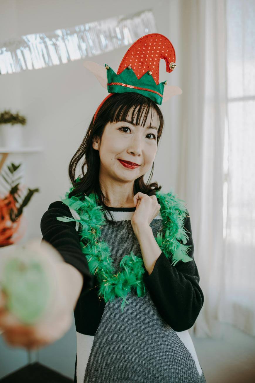A woman wearing a festive elf hat and a green feather boa smiles at the camera while holding out a decorated cookie. She stands indoors with light streaming in from a window behind her.