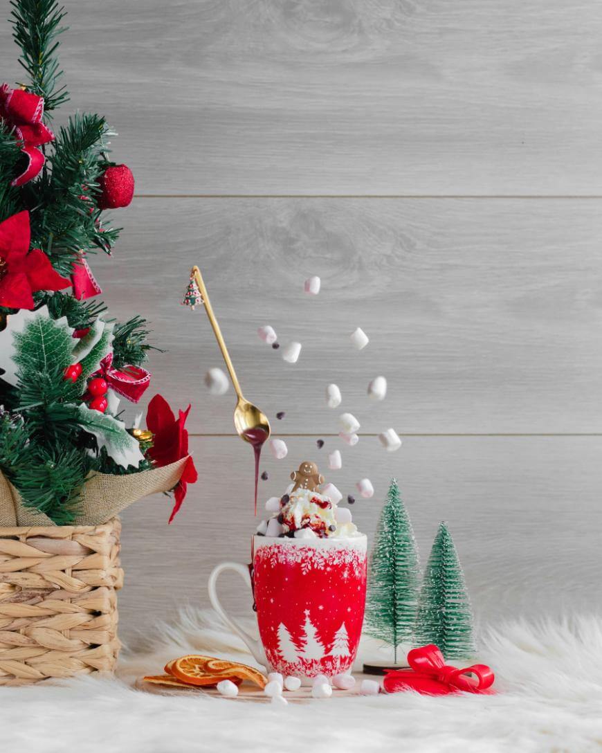 A festive red mug with white trees is topped with whipped cream, marshmallows, and gingerbread; a spoon drizzles chocolate above. A Christmas tree basket, mini trees, and orange slices sit nearby on a fluffy white surface.