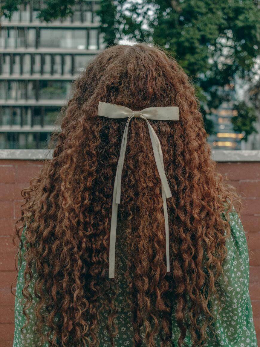 A person with long, curly brown hair, tied with a light ribbon bow, stands outdoors facing away. They wear a green top with a white floral pattern. A brick wall and blurred buildings are in the background.