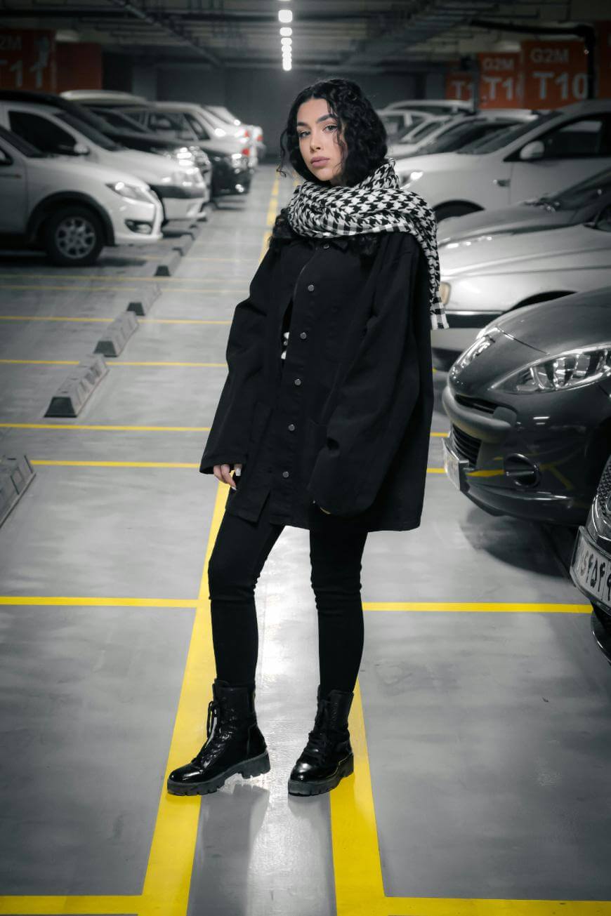 A person with curly dark hair, wearing a black coat, black pants, black boots, and a black-and-white checkered scarf, stands in a parking garage between parked cars, looking at the camera.
