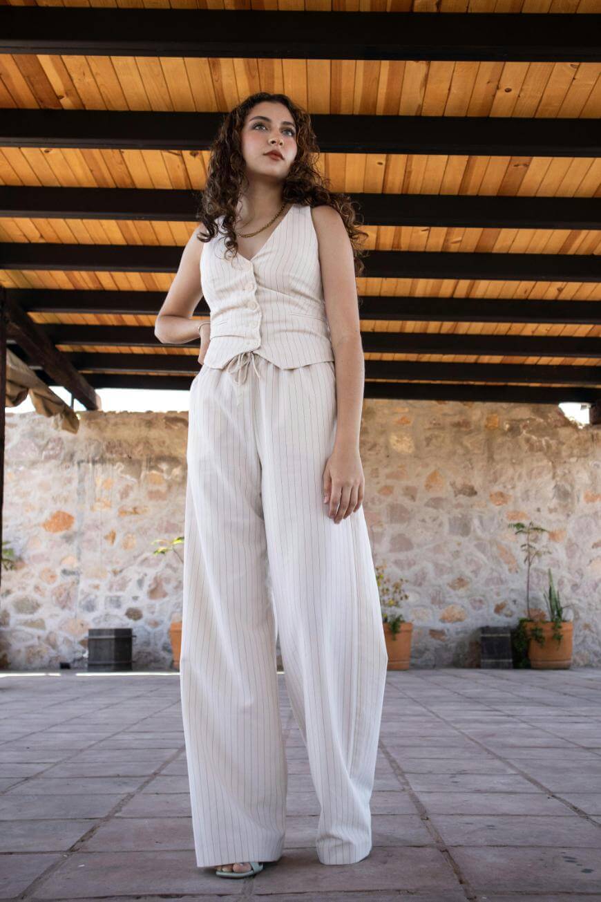 A young woman with curly hair stands confidently under a wooden roof. She wears a sleeveless, cream-colored wrap top and matching wide-leg pants with subtle stripes. Stone wall and potted plants are visible in the background.