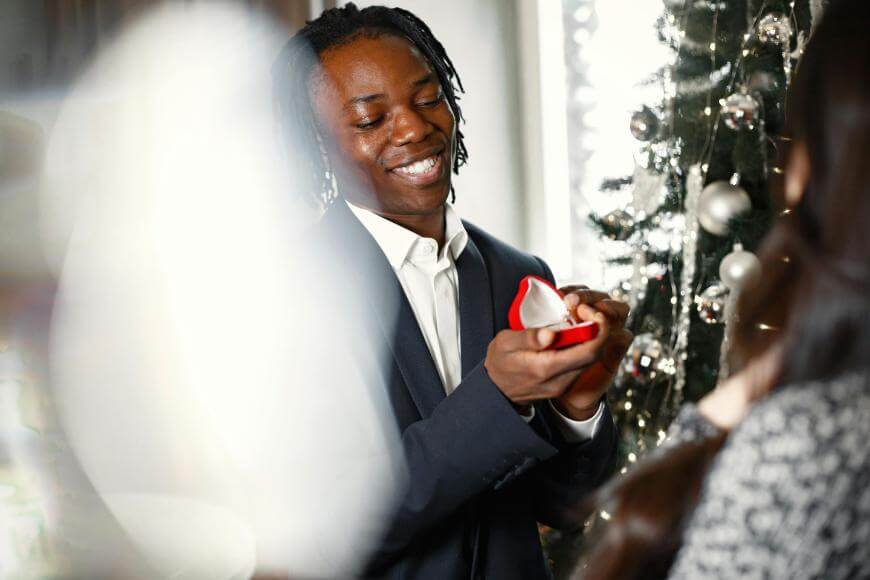 A man in a suit smiles while holding an open ring box, proposing beside a decorated Christmas tree. A person with long hair faces him, partially visible in the foreground.