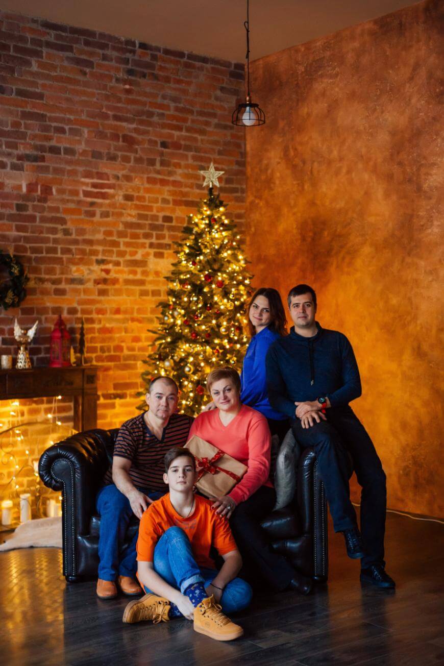 Five people pose in a cozy living room with exposed brick walls, a decorated Christmas tree, and warm lighting. Three adults sit on a black couch; one adult and a child sit in front, all smiling at the camera.