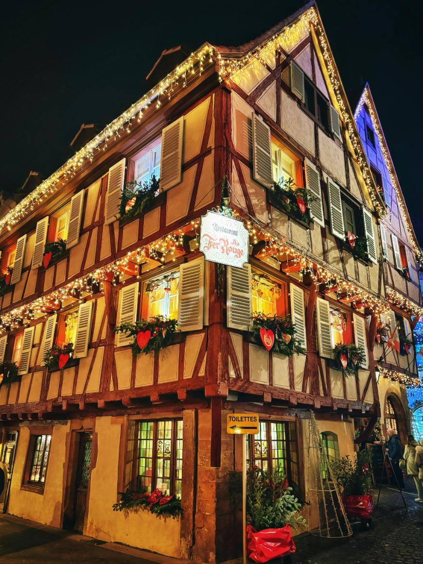 A charming, half-timbered building decorated with Christmas lights, red bows, and greenery at night. A sign reads Restaurant Aux Armes above the entrance, and a Toilettes sign is visible on the corner.