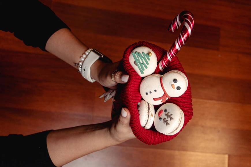 A person holds a red knit basket filled with decorated macarons shaped like snowmen, a Christmas tree, and a reindeer, along with a striped candy cane, over a wooden floor.