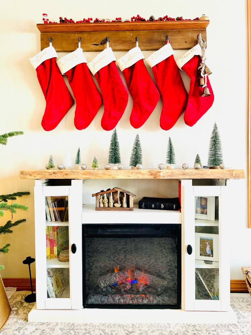A white fireplace with a wooden mantel is decorated with six red Christmas stockings, small pine trees, ornaments, and a nativity scene. A string of bells hangs from the mantel, and a cozy electric fire glows below.