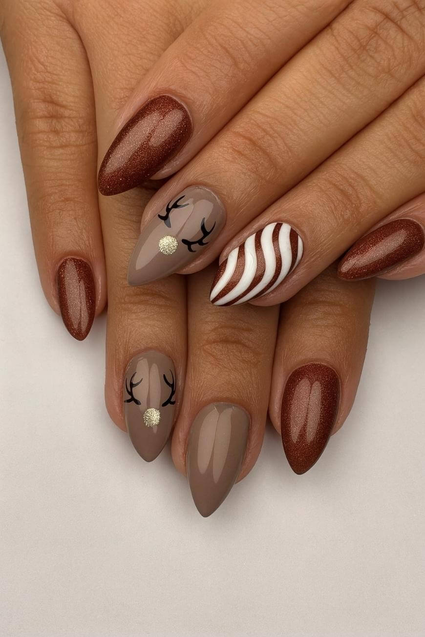 A hand with almond-shaped nails painted in brown and nude shades, featuring reindeer face art on two nails and white wavy stripes on one accent nail. The other nails have a glossy brown finish.