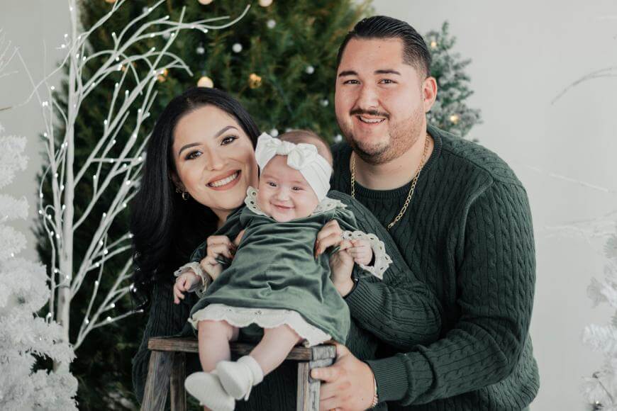 A smiling couple poses with their baby daughter in front of a decorated Christmas tree. All three are wearing matching green outfits, and the baby has a white headband and dress with lace trim. The scene is festive and cheerful.