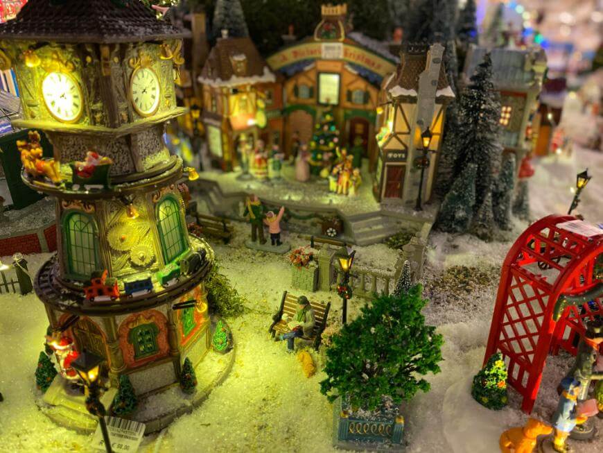 A brightly lit miniature Christmas village scene features a clock tower, festive buildings, a Christmas tree, tiny figurines, snowy ground, and a red bridge, all decorated for the holiday season.