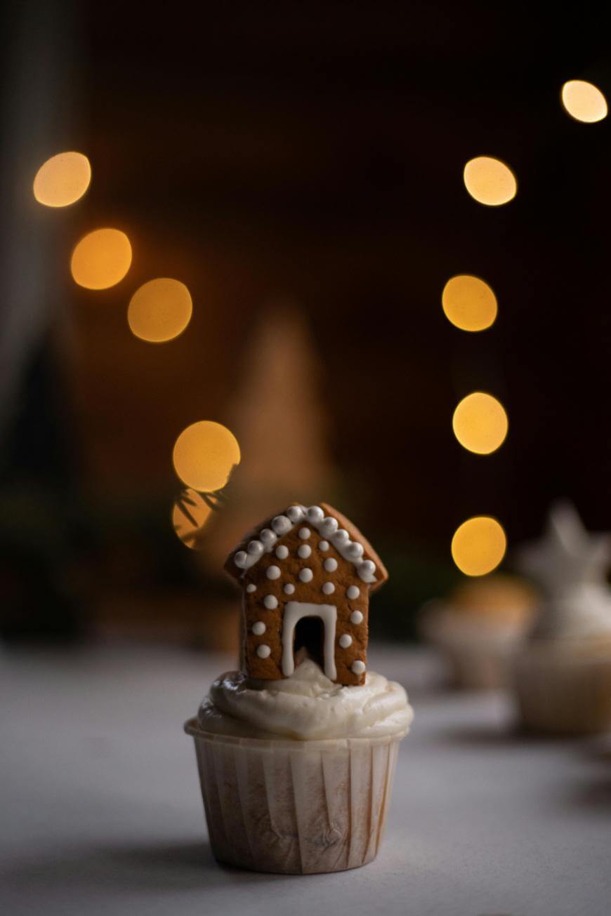 A cupcake topped with a small gingerbread house, decorated with white icing, sits on a white surface. Warm, blurred yellow lights glow softly in the dark background.