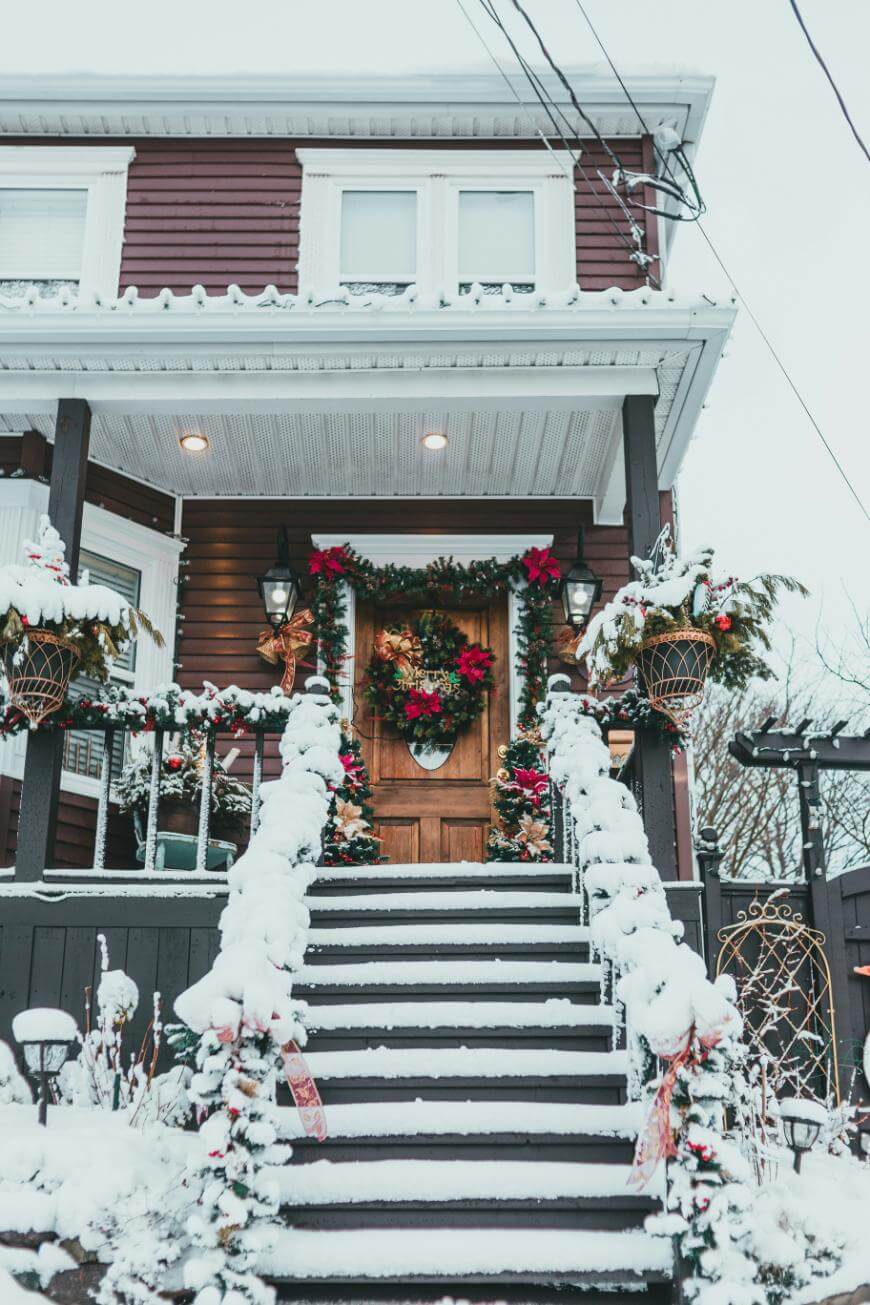 A wooden front door decorated with a Christmas wreath, stairs and railings covered in snow, and festive garlands and red bows on a house exterior during winter.